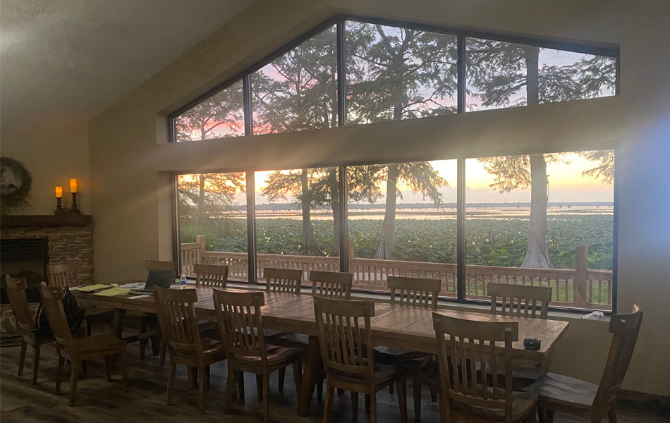 an extra large dinning table in front of a floor-to-ceiling window with a lake view