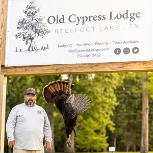 Man standing with a turkey in front of the Old Cypress Lodge sign